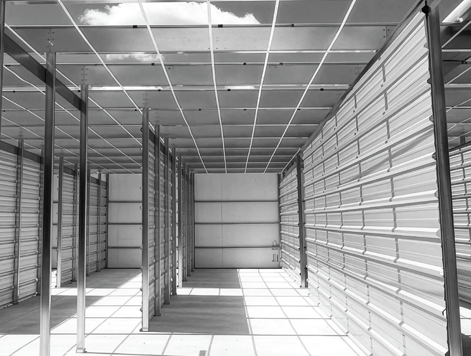 A black and white photo of the interior of a metal storage facility under construction, showing corrugated partition walls, a concrete floor, and an open ceiling grid casting geometric shadows.