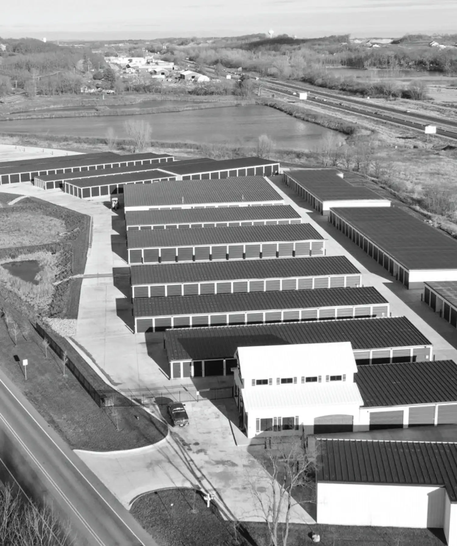 A black and white aerial view of a large self-storage facility featuring multiple rows of long storage buildings, paved driveways, and a white office building in the foreground, situated near a highway and a body of water.
