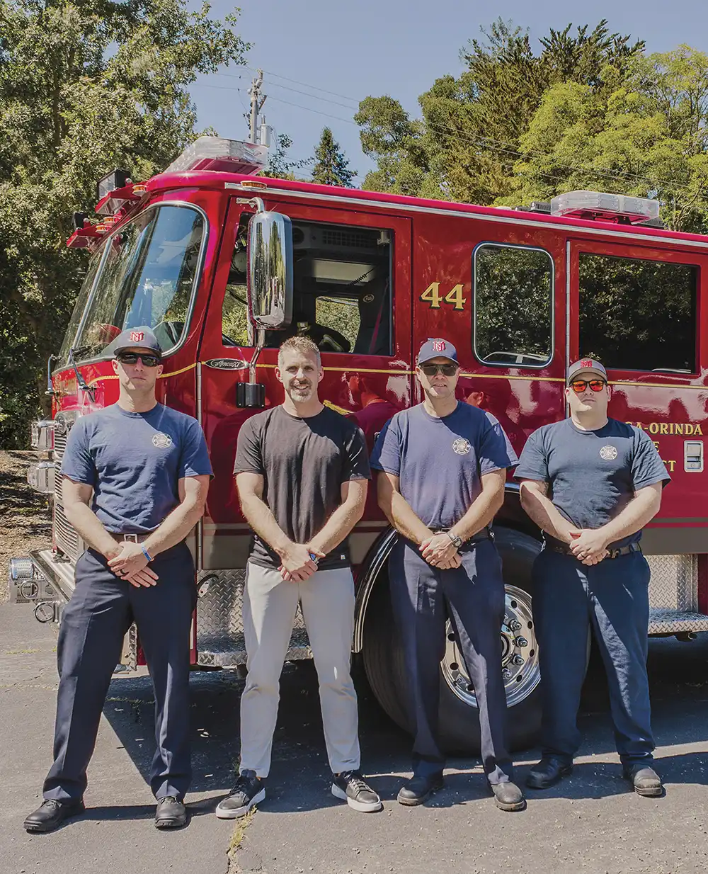 Andrew Hess, wearing civilian clothes, stands in a line with three uniformed firefighters in front of a parked red Moraga-Orinda Fire District firetruck.