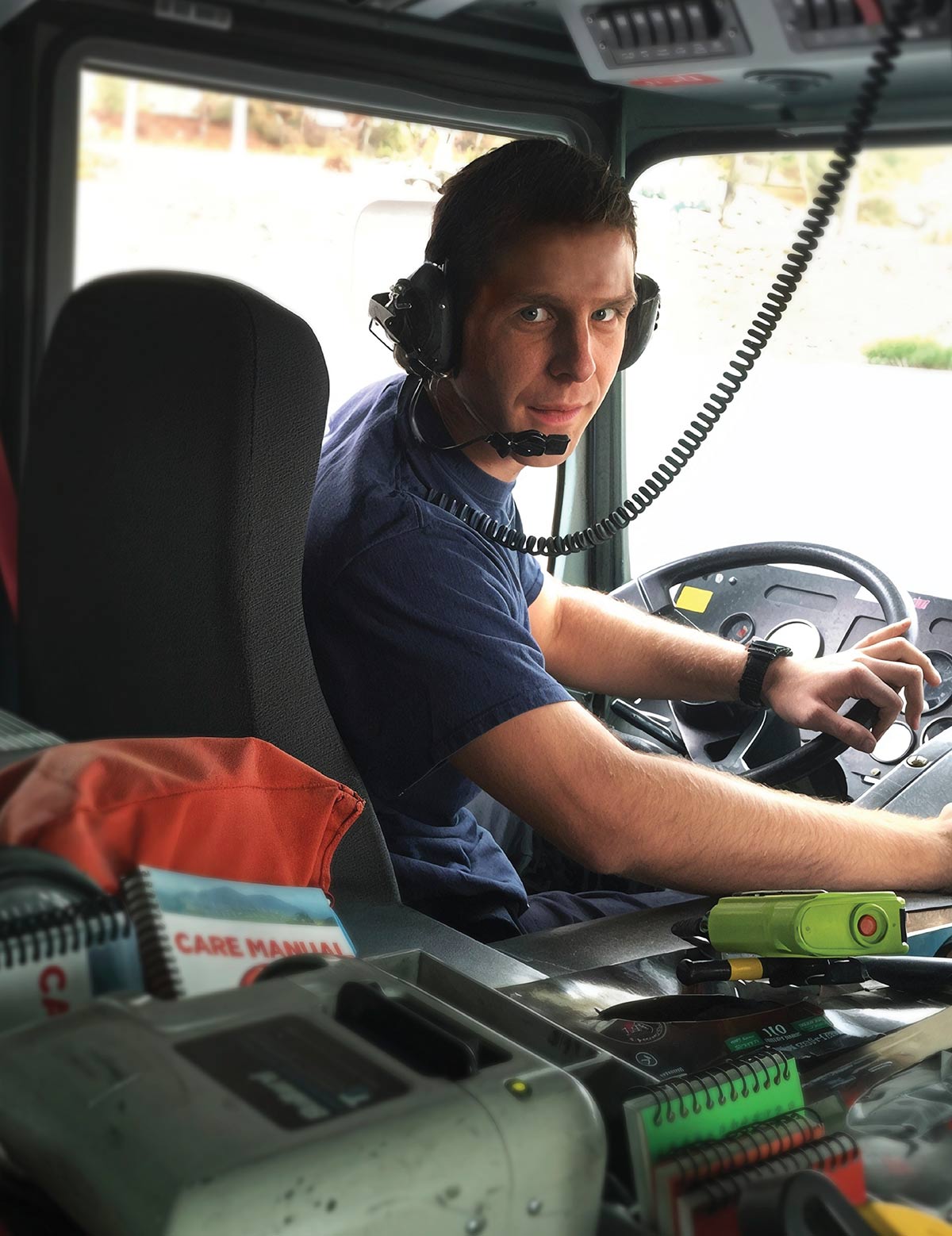 Andrew Hess wears a headset while sitting in the driver's seat of a vehicle, looking over his shoulder at the camera with his hands on the steering wheel.