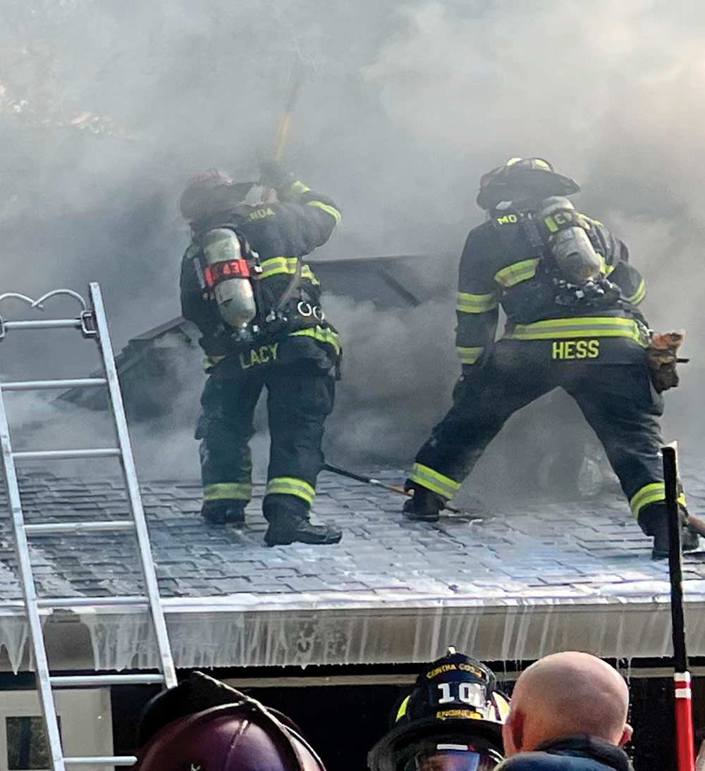 Andrew Hess and another firefighter in full turnout gear stand on a shingled roof surrounded by thick gray smoke, using tools to ventilate the roof during a fire.