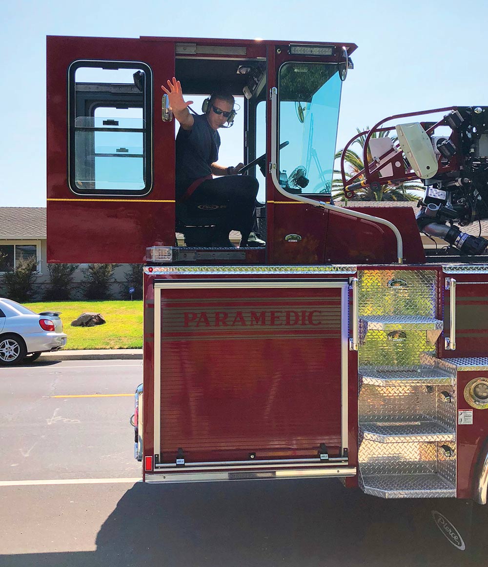 Andrew Hess wearing sunglasses and a headset sits in the driver's seat of a red fire engine, waving his right hand out the open door. The side of the truck displays the word “PARAMEDIC”.