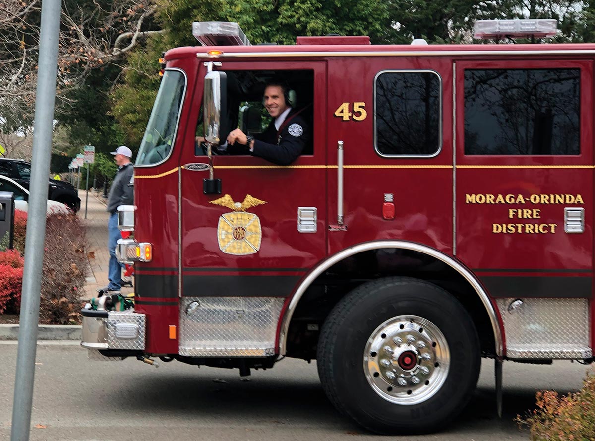 Andrew Hess smiles out the driver's side window of a red Moraga-Orinda Fire District firetruck. The truck door features the number 45 and a gold district emblem.