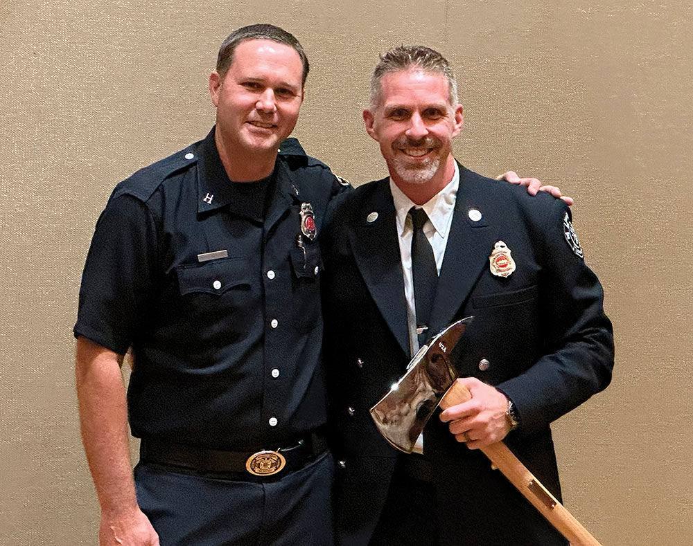 Andrew Hess and Brad Nygard stand together smiling in dark fire department uniforms. Nygard wears a short-sleeve uniform shirt while Hess wears a dress uniform jacket and holds a silver ceremonial axe.