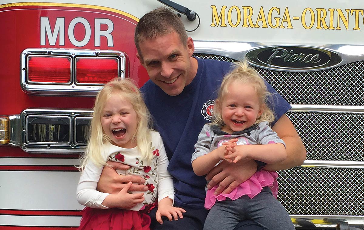 Andrew Hess sits in front of the grill of a red Moraga-Orinda firetruck, smiling while holding two young, laughing girls on his lap.
