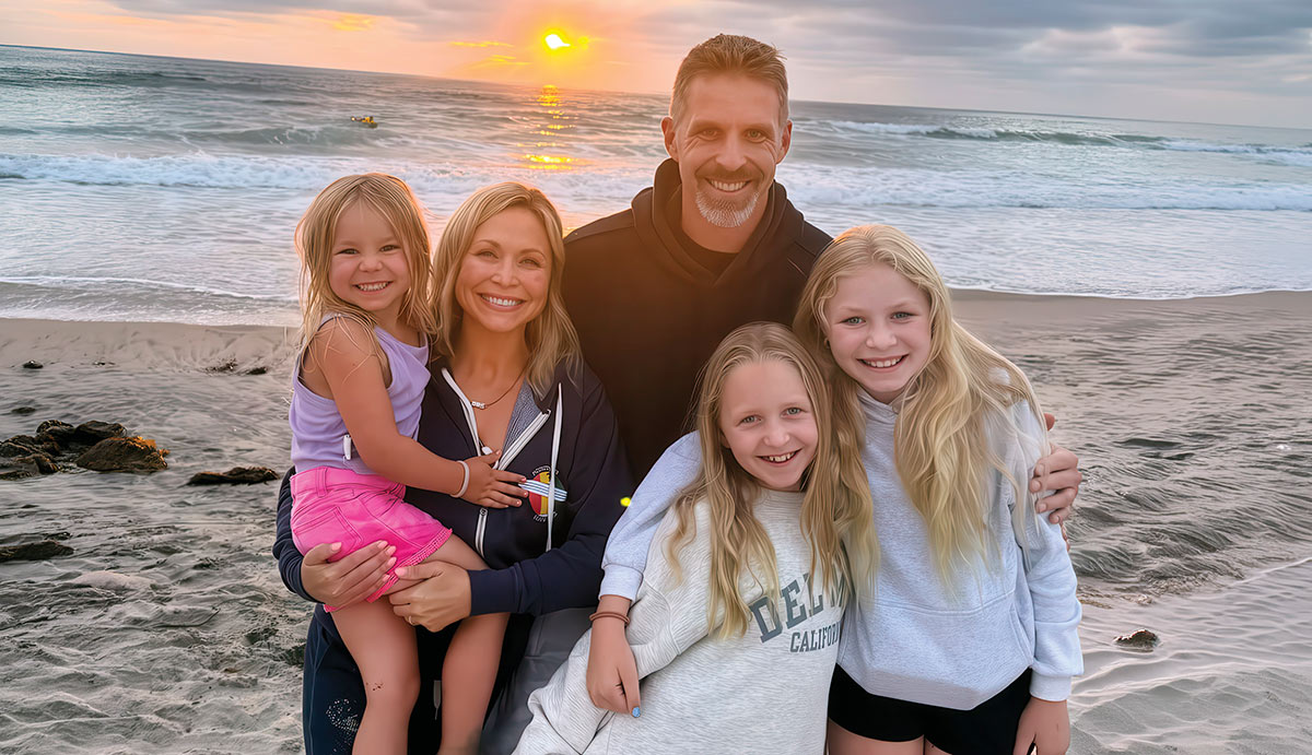 Andrew Hess, his wife, and their three daughters smile together while posing on a sandy beach. The ocean and a sunset are visible in the background behind them.