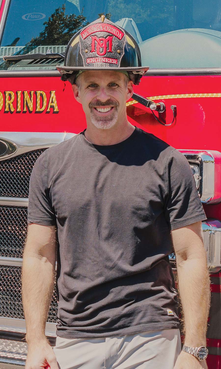 Andrew Hess smiles while standing in front of a red firetruck. He is wearing a black t-shirt and a black firefighter's helmet with a front shield that reads "Moraga-Orinda Engineer Hess".