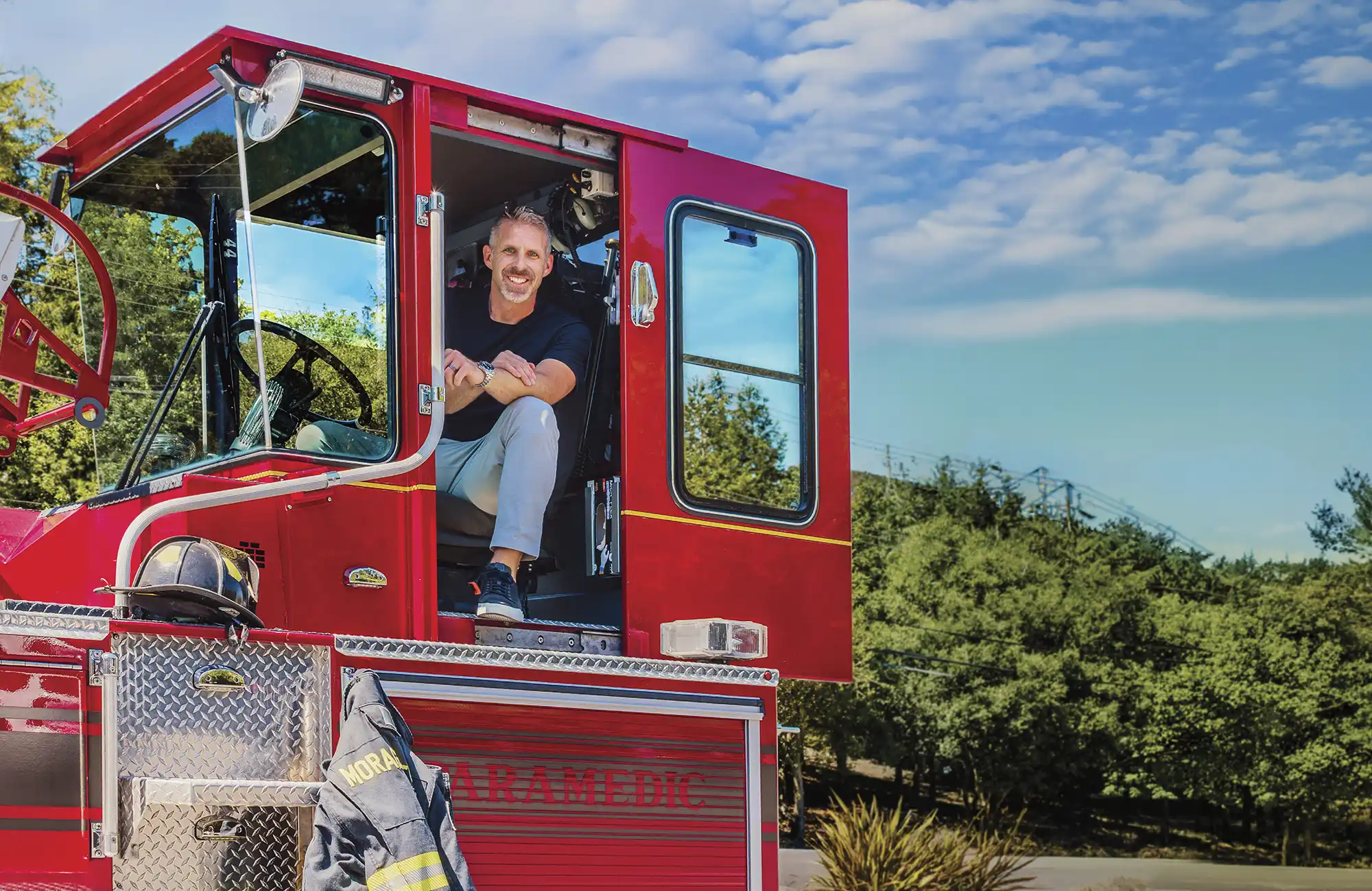 Andrew Hess sits in the open driver-side door of a parked red firetruck on a sunny day, smiling and leaning out with his arms crossed. A firefighter's coat and helmet hang on the side of the truck.