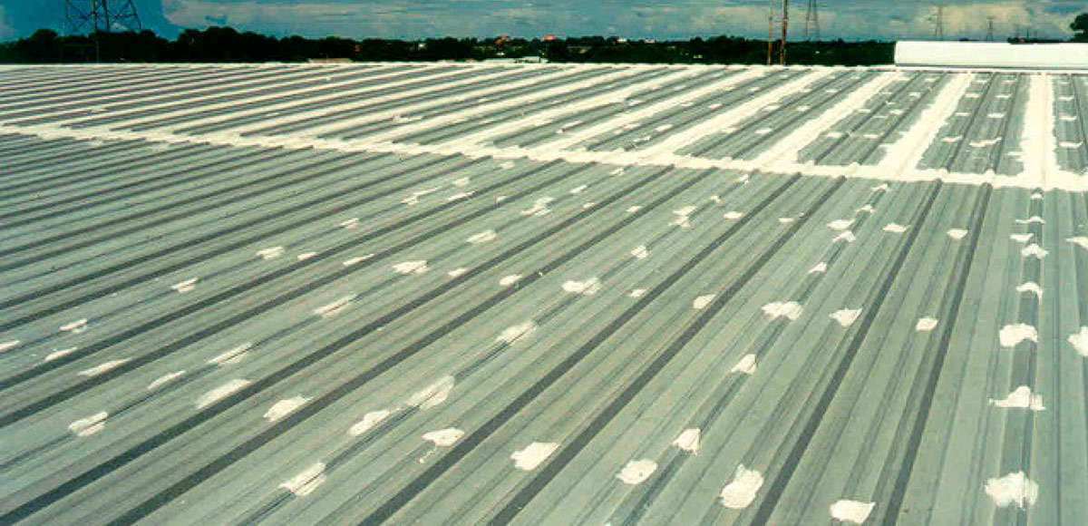 Wide view of a large corrugated metal building roof covered in numerous small, white patches of sealant over the seams and rows of fasteners.
