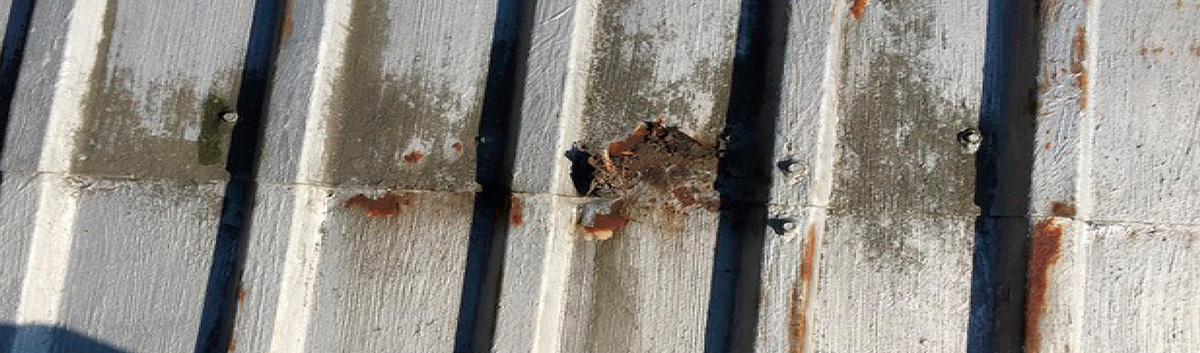 Close-up of overlapping corrugated metal panels where a patch of gray protective coating has broken away, revealing dark brown rust and corrosion on the metal surface underneath.
