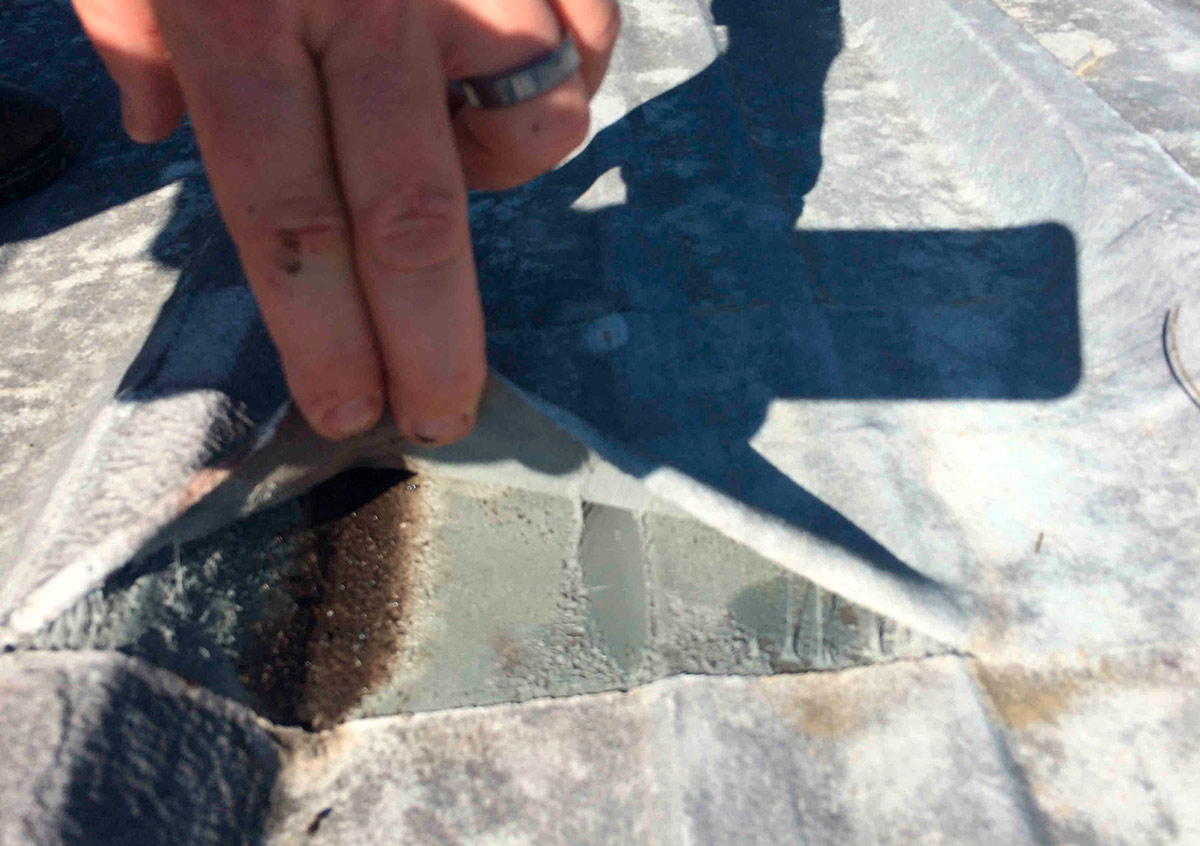 A hand peels back a layer of flexible gray roof coating from a weathered metal roof panel, exposing a dark patch of rust and moisture beneath it.