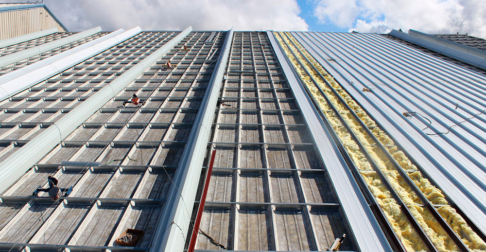 Low-angle view of a large roof retrofit in progress, showing a metal grid framework over old panels, exposed yellow foam insulation, and newly installed white corrugated roofing panels.