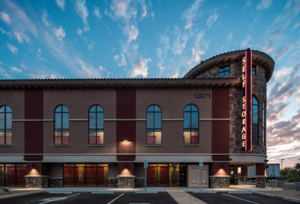 Exterior of a multi-story self-storage facility with a stone tower corner and neon "Self Storage" signage at dusk.