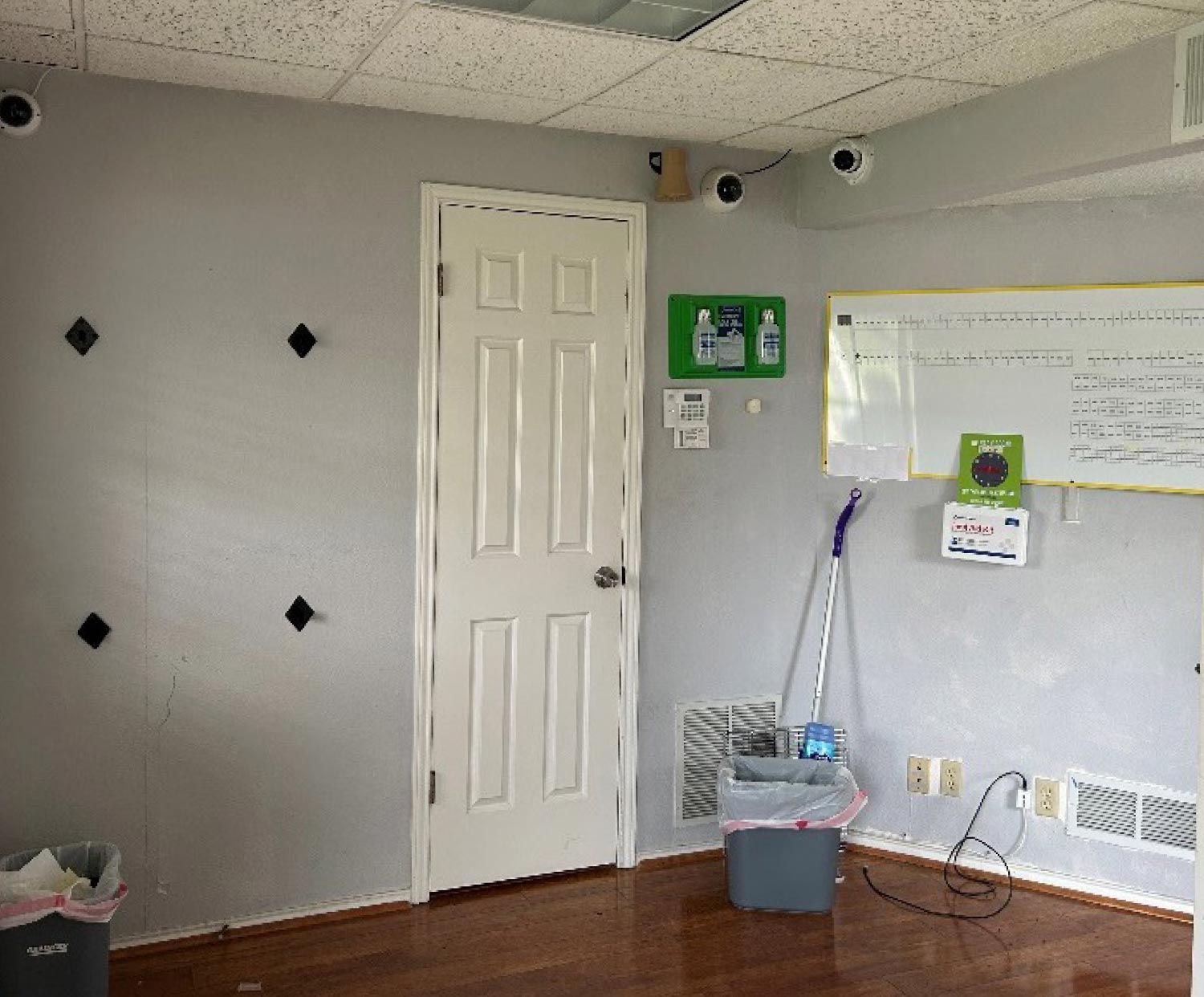 An outdated office room before renovation, featuring plain gray walls, a whiteboard, security cameras, and cleaning supplies.