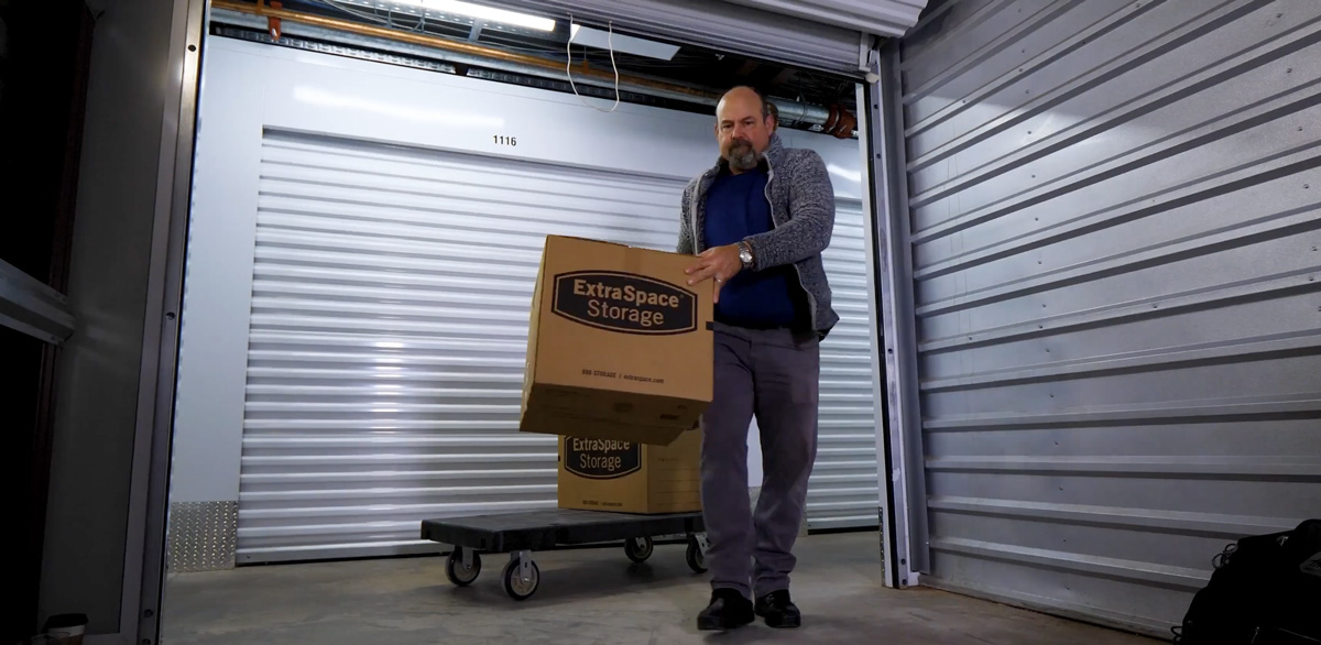 Man carrying an Extra Space Storage cardboard box inside a indoor storage unit hallway near a flatbed cart.