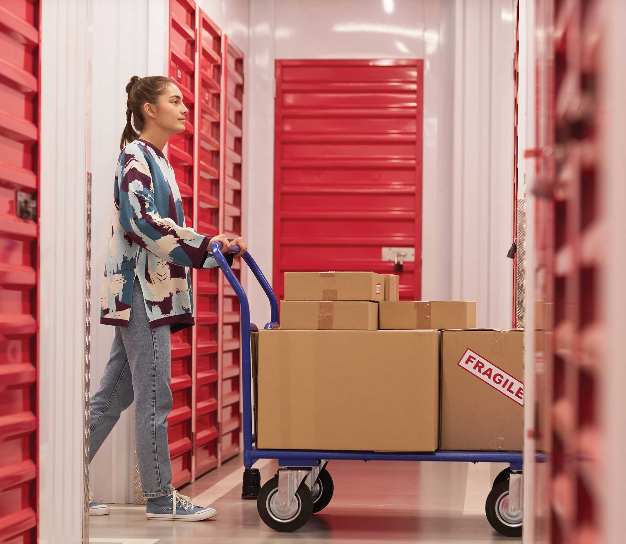 Woman pushing a box-filled cart down a self-storage corridor with red doors.