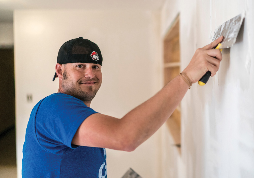 A man wearing a baseball cap smiles while applying joint compound to a wall.