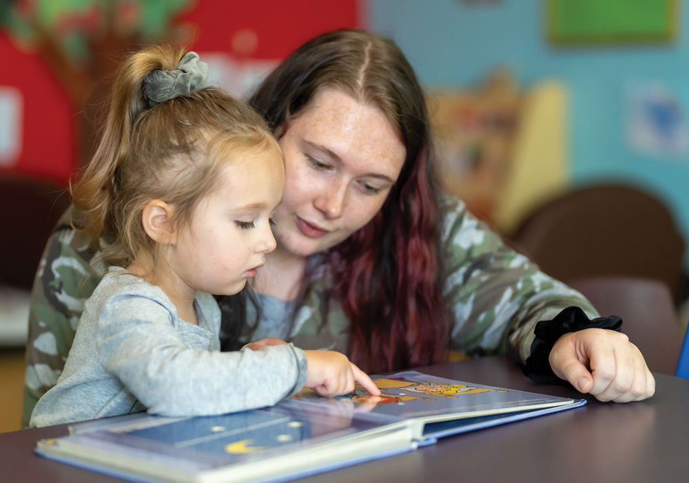 A woman with long hair reads a picture book to a young child.