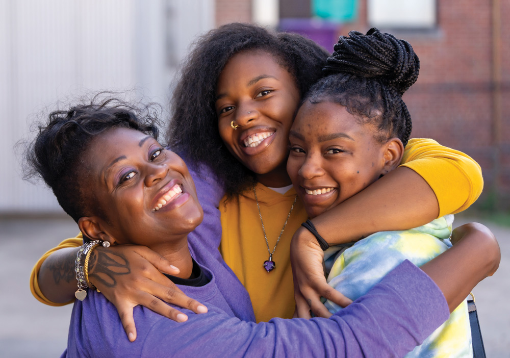 Three women embrace and smile warmly together in an outdoor setting.