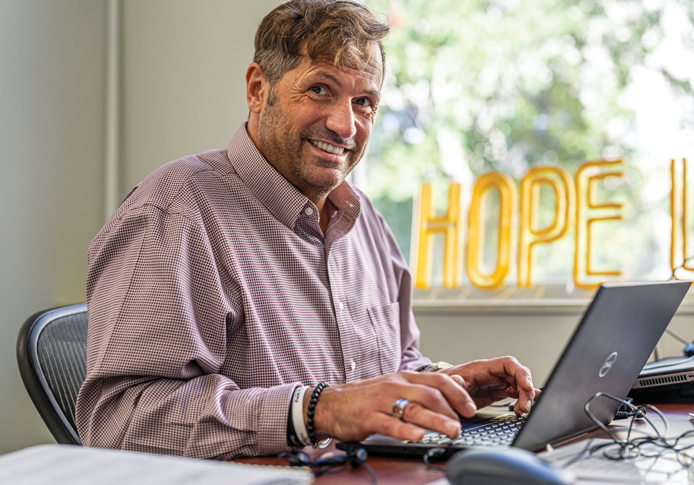 A man smiles while working on a laptop at a wooden desk.