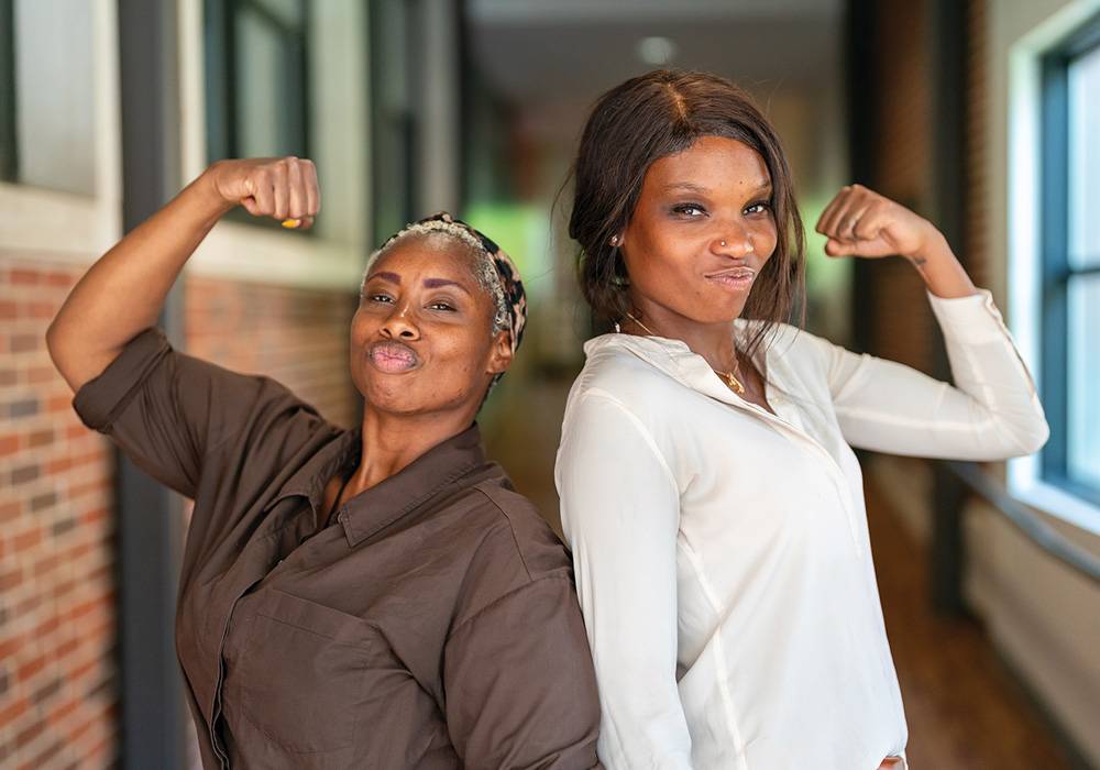 Two women standing in a hallway flex their biceps while posing for a photo.