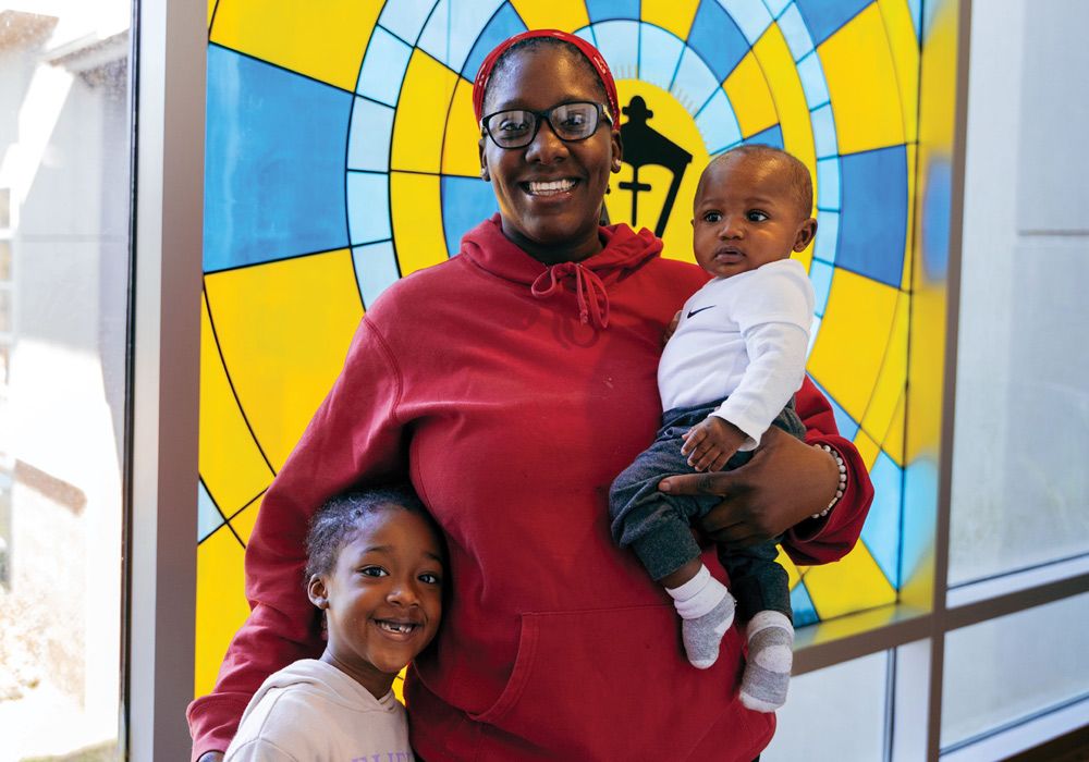 A smiling woman stands with two children in front of a colorful stained-glass window.
