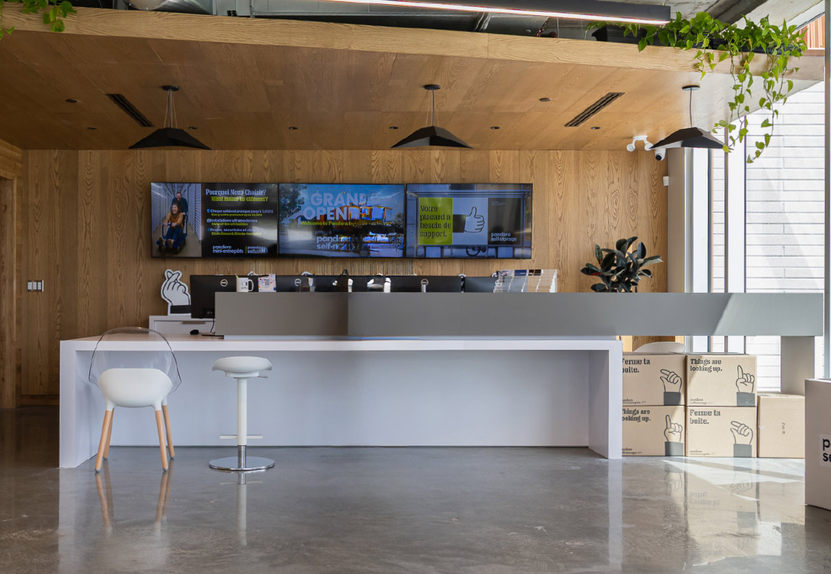Modern reception desk at Pandora Mini-Entrepôts featuring wood-paneled walls, hanging lights, and wall monitors.