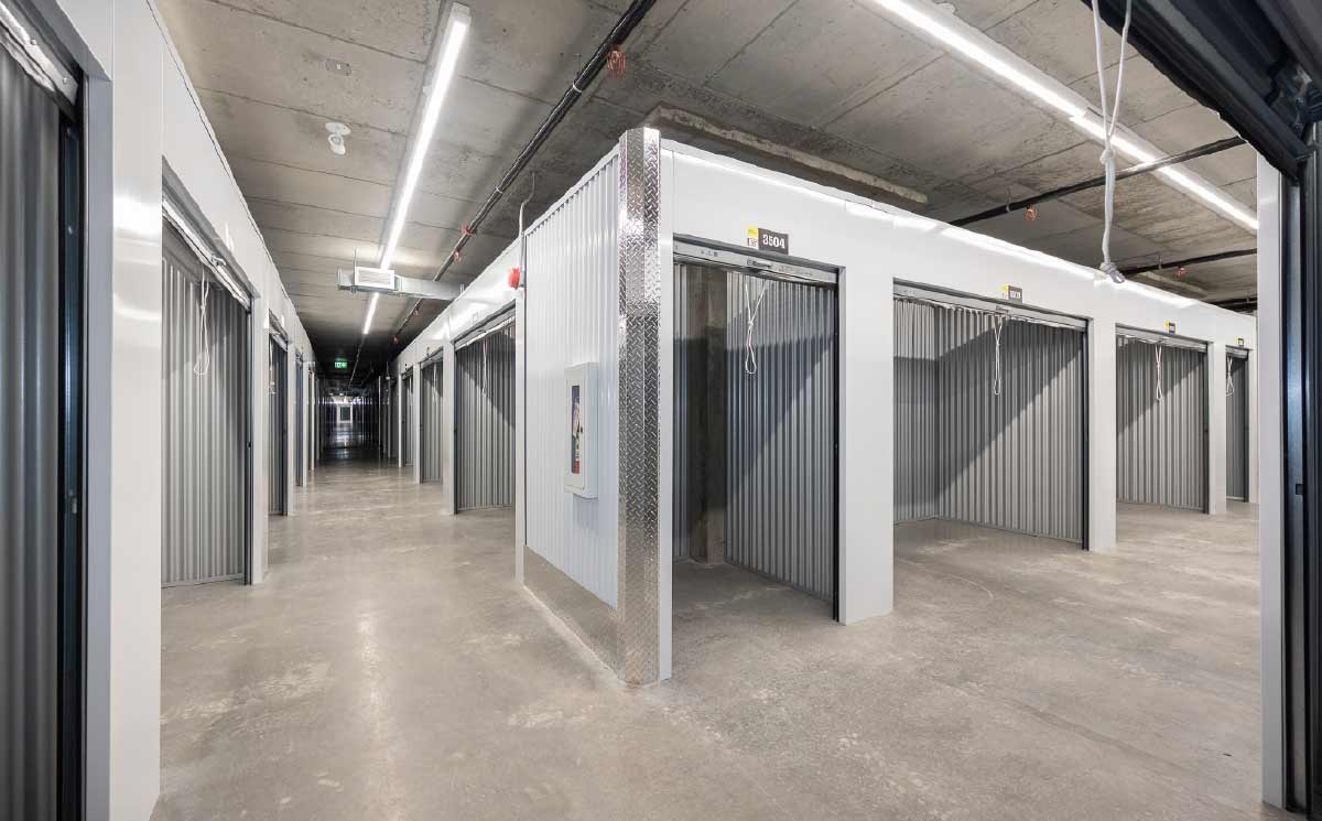 Well-lit indoor hallway lined with white, clean self-storage units with roll-up metal doors.