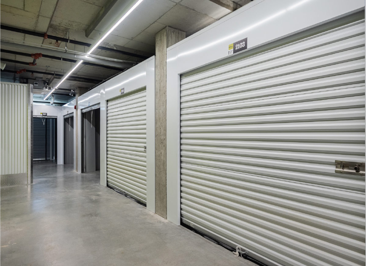 Well-lit indoor hallway lined with white, clean self-storage units with roll-up metal doors.