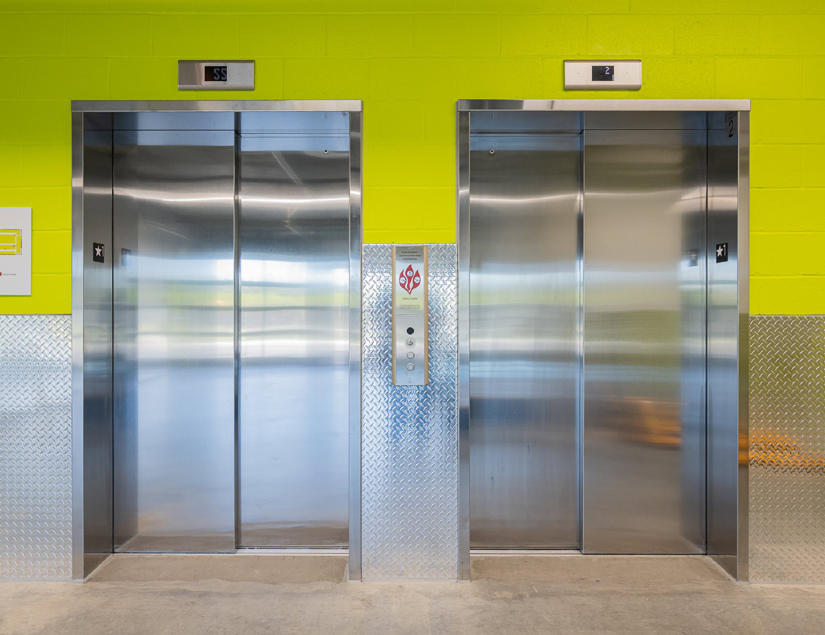 Two stainless steel elevator doors set against a lime-green wall with diamond-plate aluminum trim.