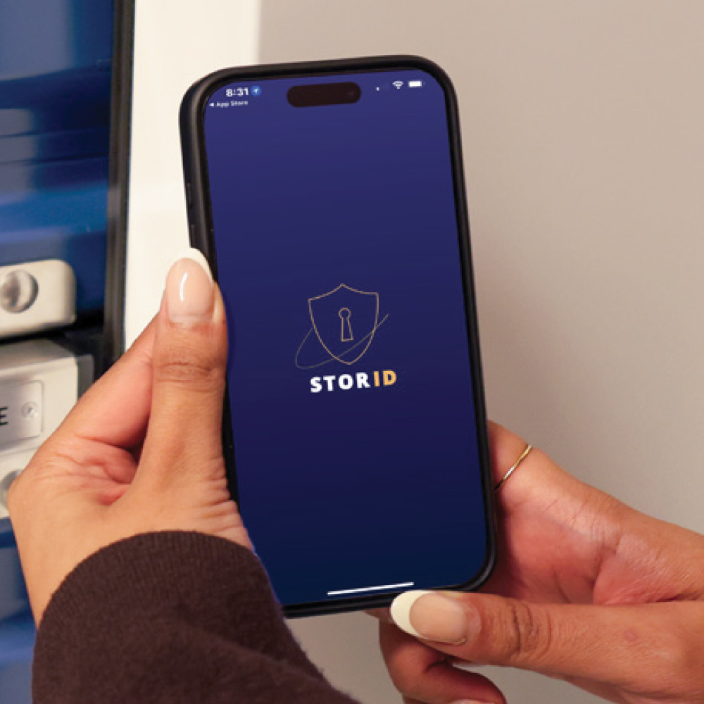 Close-up of hands holding a smartphone displaying the StorID app home screen next to an access panel.