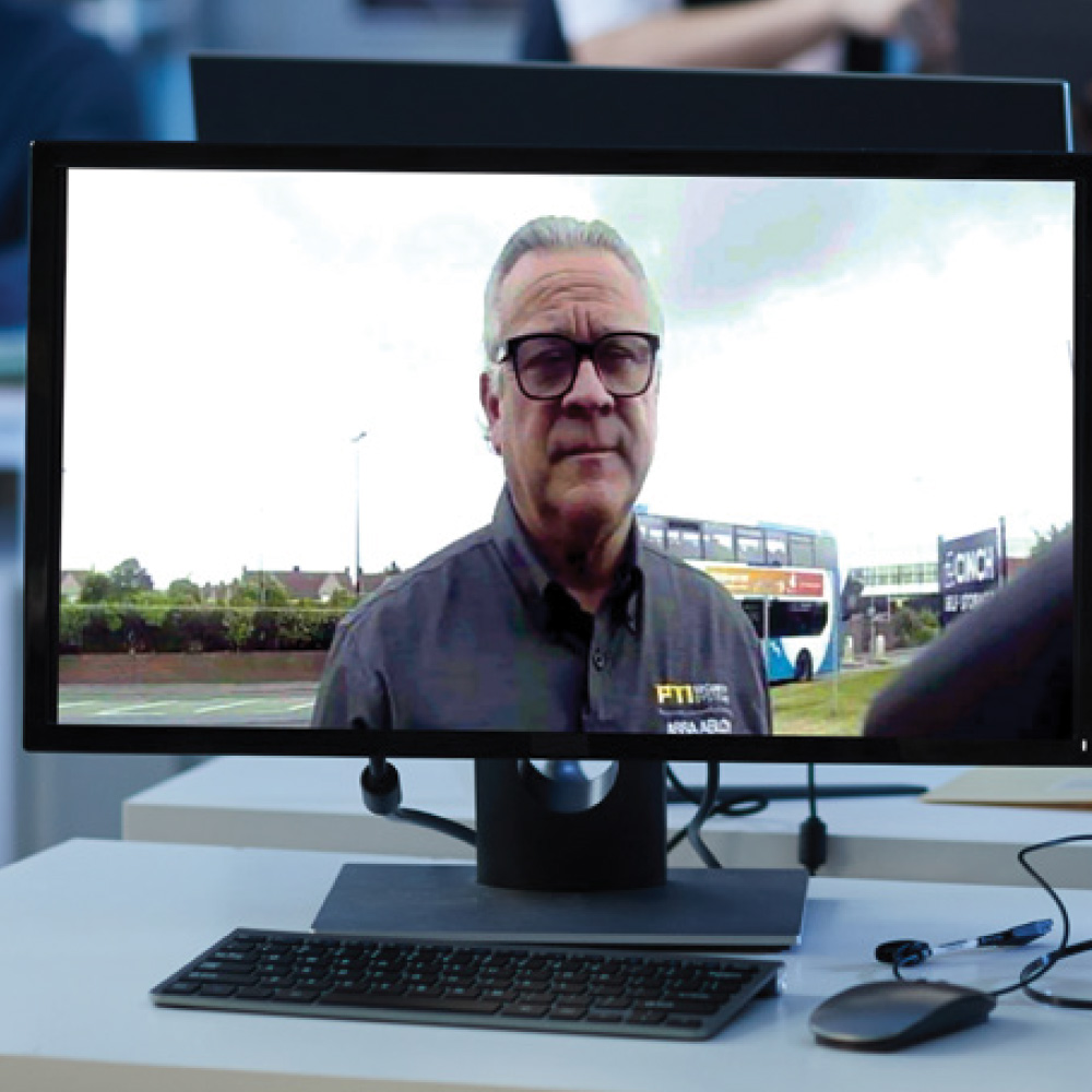 Computer monitor on an office desk displaying a video call with a man wearing glasses and a branded PTI shirt.