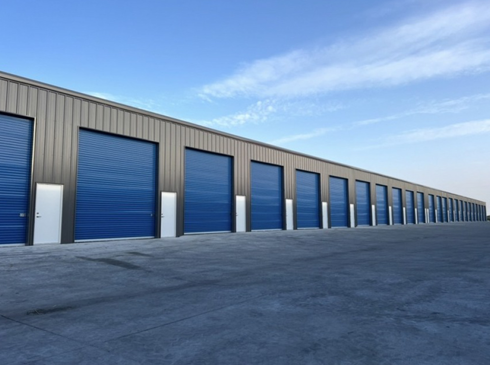 Long row of dark metal self-storage facility buildings with blue garage roll-up doors and white entry doors.