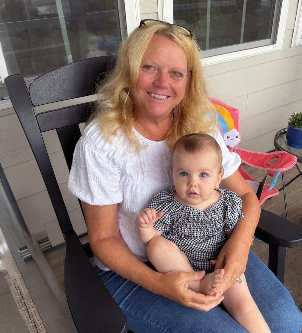 Smiling woman sitting in a dark wooden chair holding a baby in a checkered outfit on her lap.