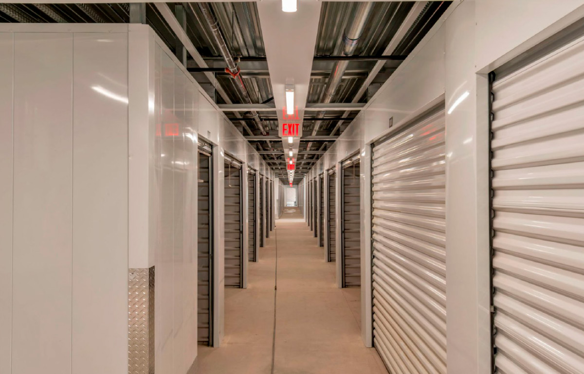 Bright, clean interior hallway of a self-storage facility featuring white roll-up storage unit doors and exit signage.
