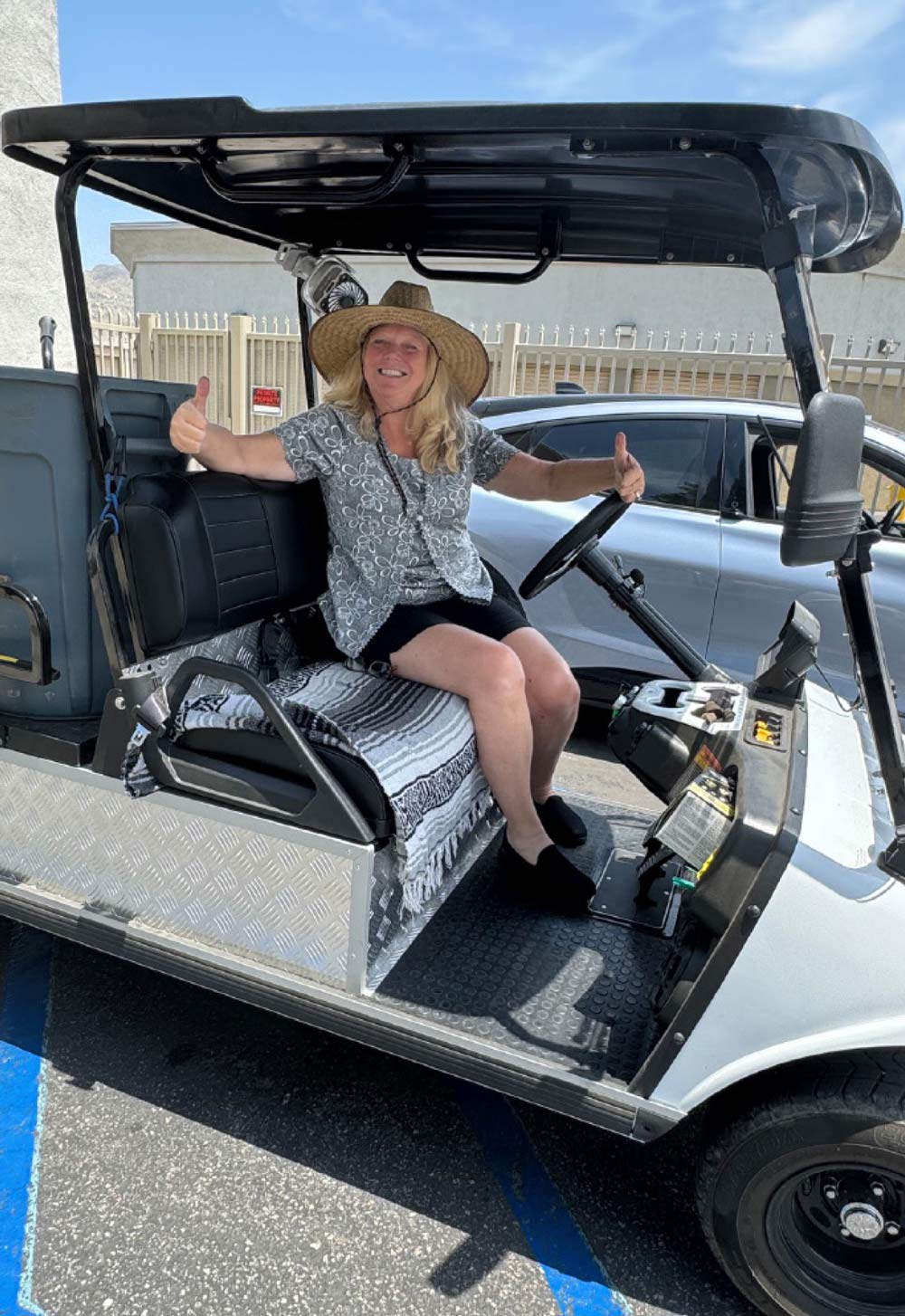 Woman wearing a straw hat giving two thumbs-up while sitting at the wheel of a golf cart.