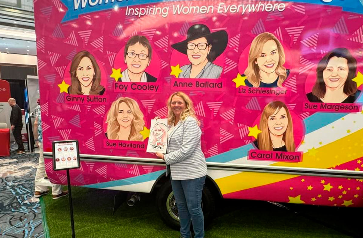 Sue Haviland posing holding a caricature drawing in front of an "Inspiring Women Everywhere" banner.