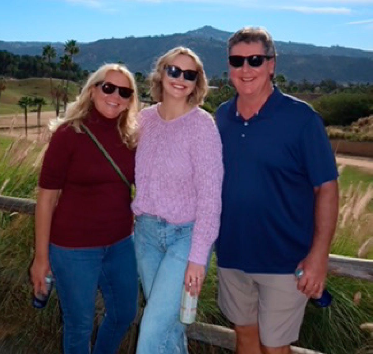 Three people wearing sunglasses posing together outdoors in front of scenic hills.