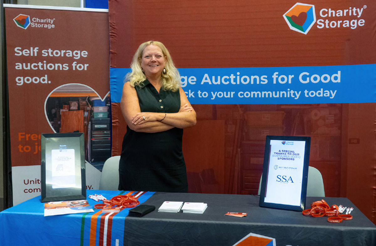 Sue Haviland posing with crossed arms behind a Charity Storage booth table at a trade show.