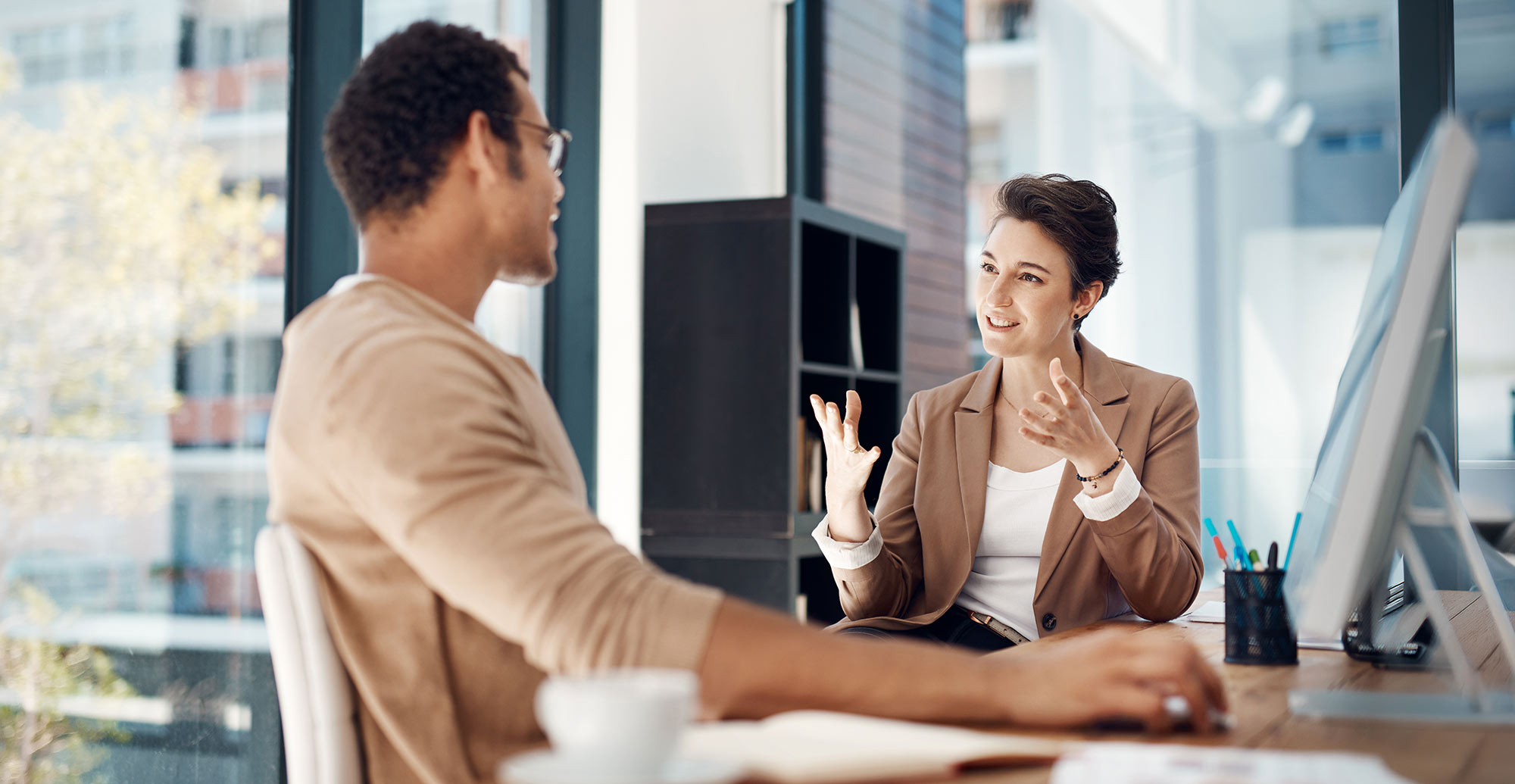 Professional woman in a blazer speaking with a colleague in a bright, modern office.