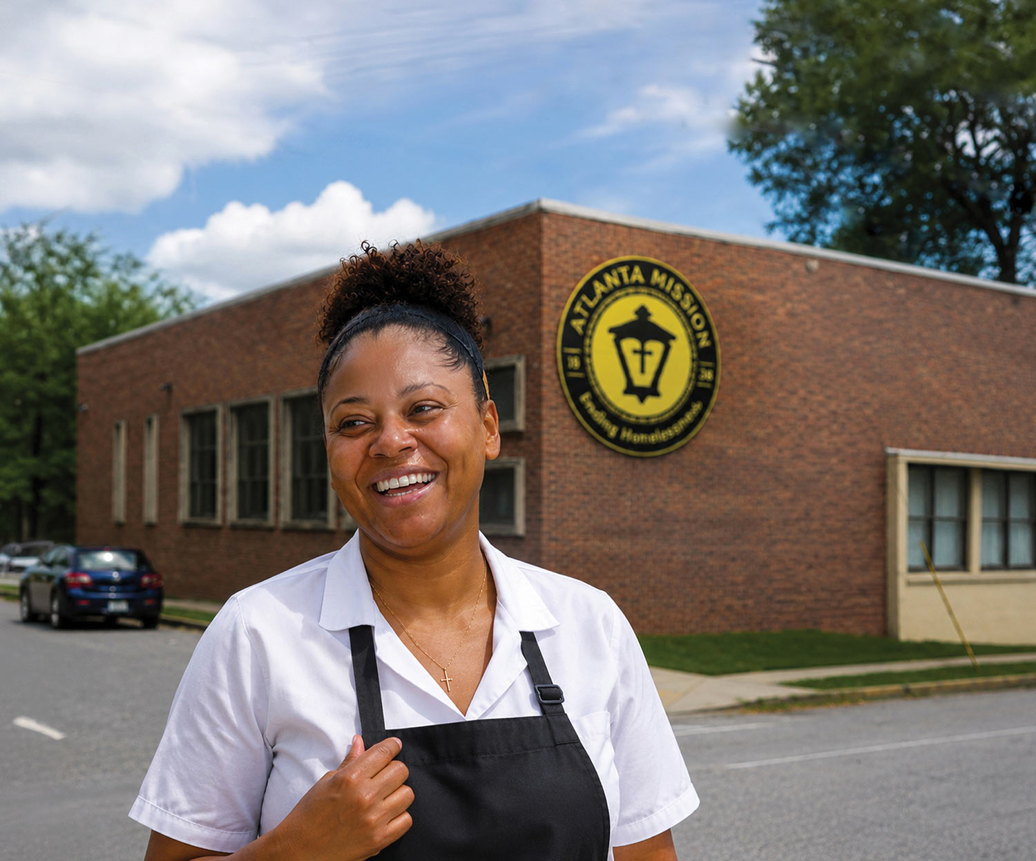 Woman in white shirt and apron smiling in front of Atlanta Mission brick building.